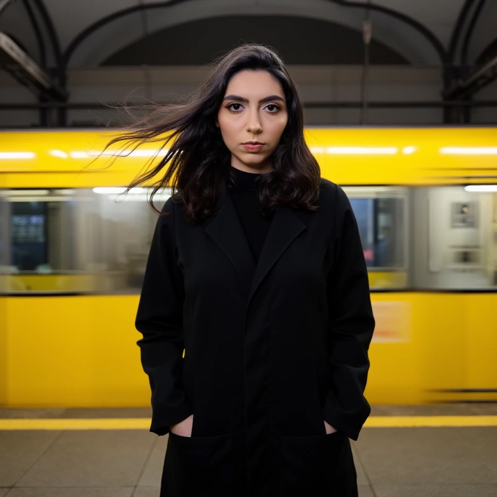 Lara Roldi em uma plataforma de metrô, com um trem amarelo em movimento ao fundo